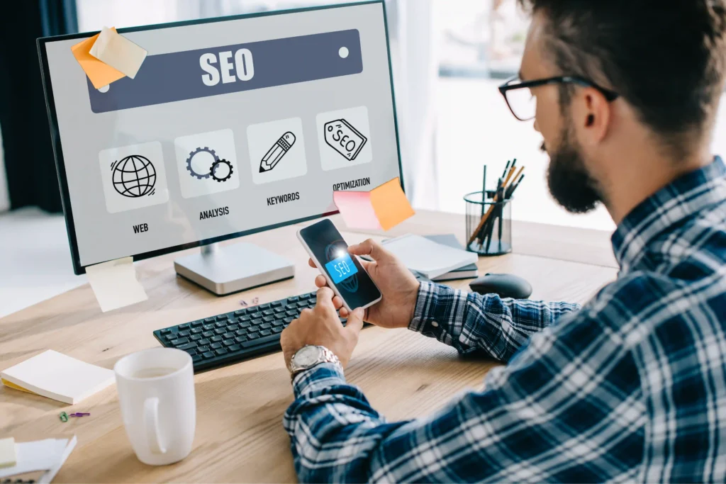 Young SEO manager analyzing technical SEO steps on a smartphone and desktop monitor in a modern workspace