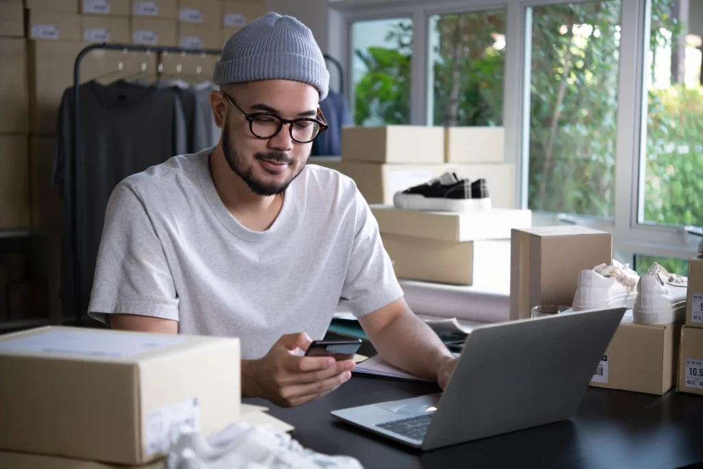 E-commerce seller using laptop and mobile phone in a shipping-ready product workspace.