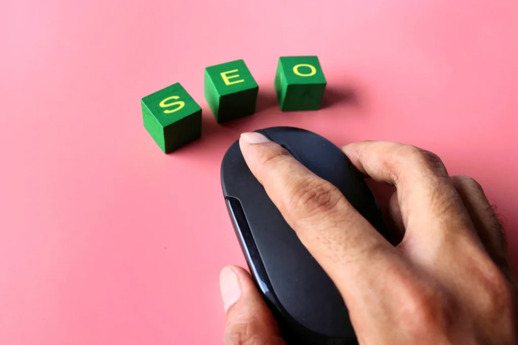 Hand clicking mouse beside green blocks spelling SEO on a pink background, symbolizing seo marketing free access.