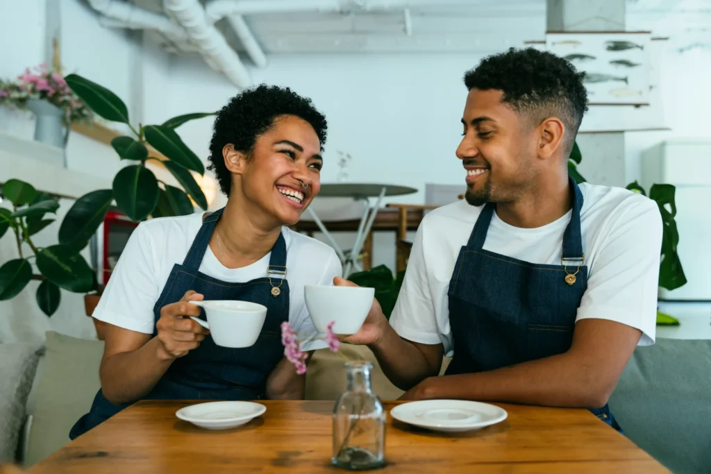 Two smiling café workers in denim aprons sharing coffee and laughter at a cozy table, symbolizing teamwork and great café culture.