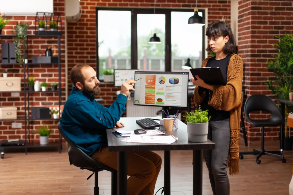 Two colleagues in a digital media marketing company reviewing campaign analytics on a computer screen in a modern office.