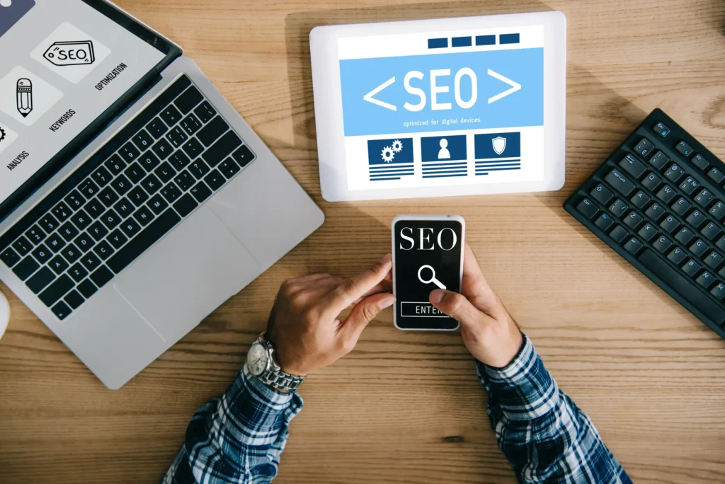 Person using mobile and laptop tools to perform SEO digital marketing services with keyword and optimization icons.