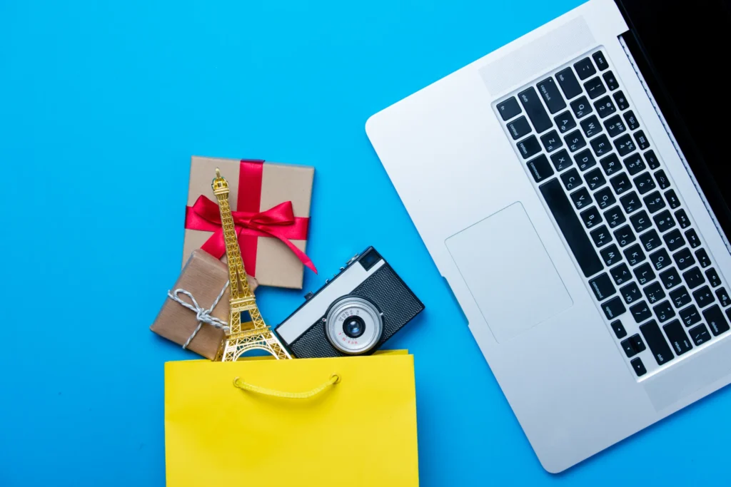 Yellow shopping bag with camera, Eiffel Tower model, and gifts beside a laptop showing the lifestyle appeal of an ecommerce branding agency.