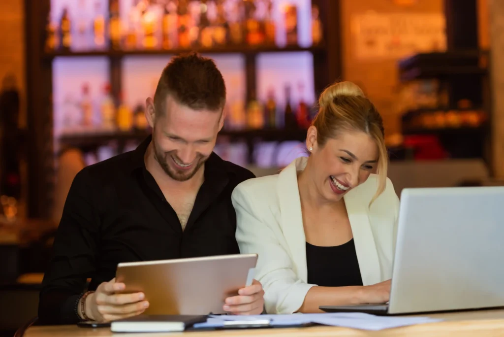 Two restaurant owners laughing while reviewing marketing strategies on a tablet and laptop, representing teamwork with a digital marketing agency for restaurants.