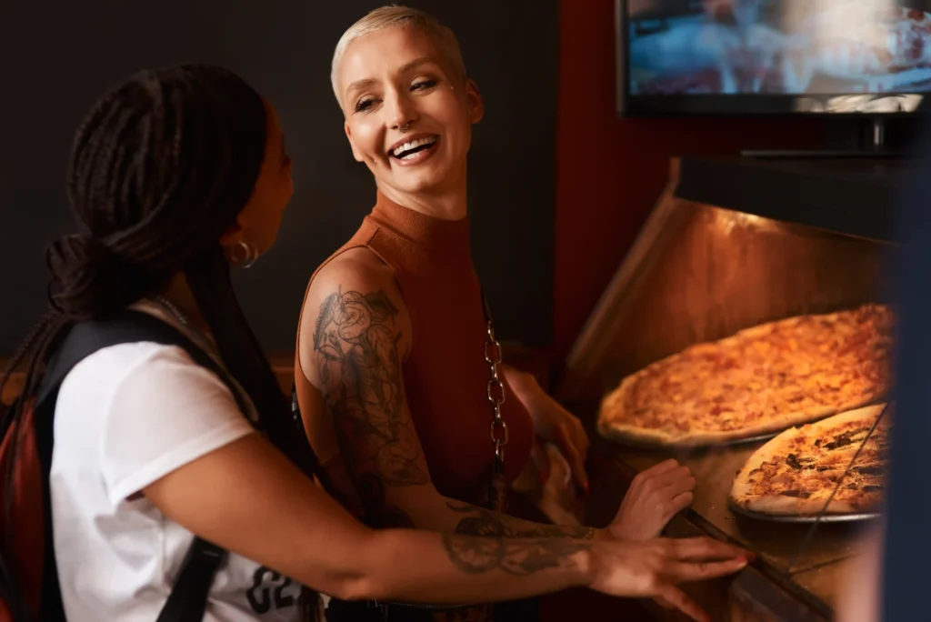 Two women smiling at a pizza counter, representing customer experience in pizza marketing ideas.