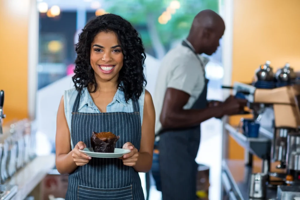 Smiling waitress holding a muffin at a café counter, reflecting hospitality and staff participation in unique restaurant promotion ideas.