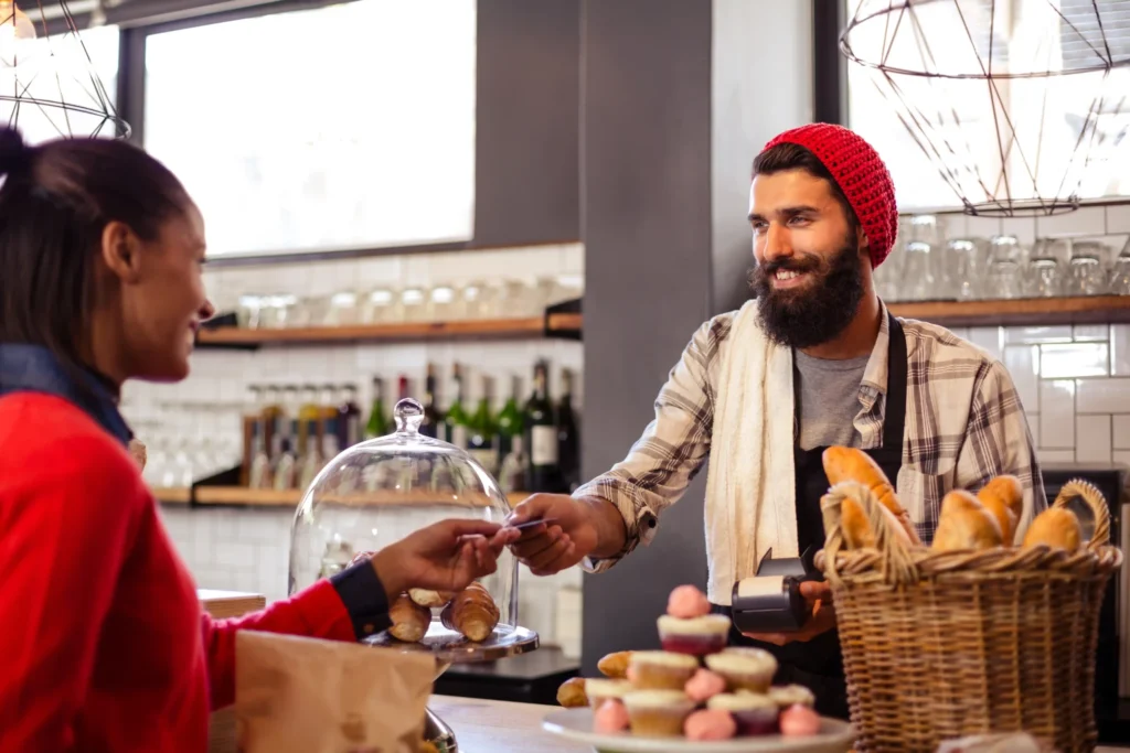 Bearded barista handing change to a customer beside pastries and bread, symbolizing loyalty-driven and in-store unique restaurant promotion ideas.