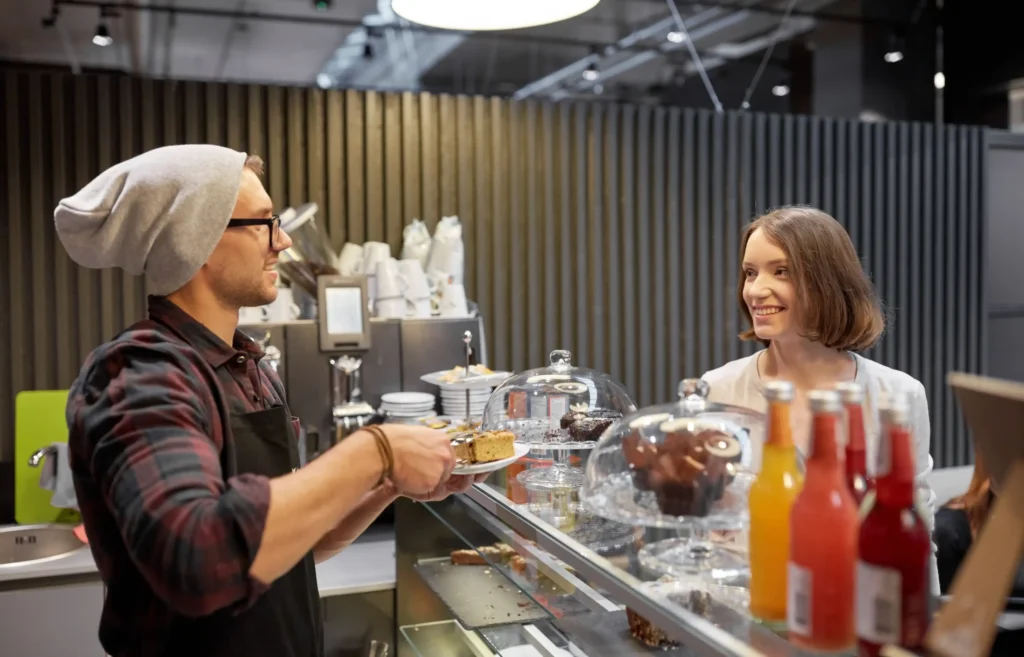 Barista serving a smiling customer at a modern café counter filled with pastries and drinks, showing friendly service.