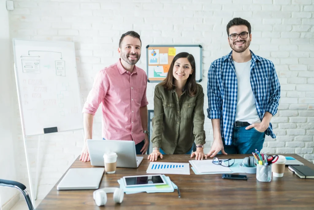Three colleagues at a web design and marketing agency standing at a desk with laptops and planning materials during a strategy session.
