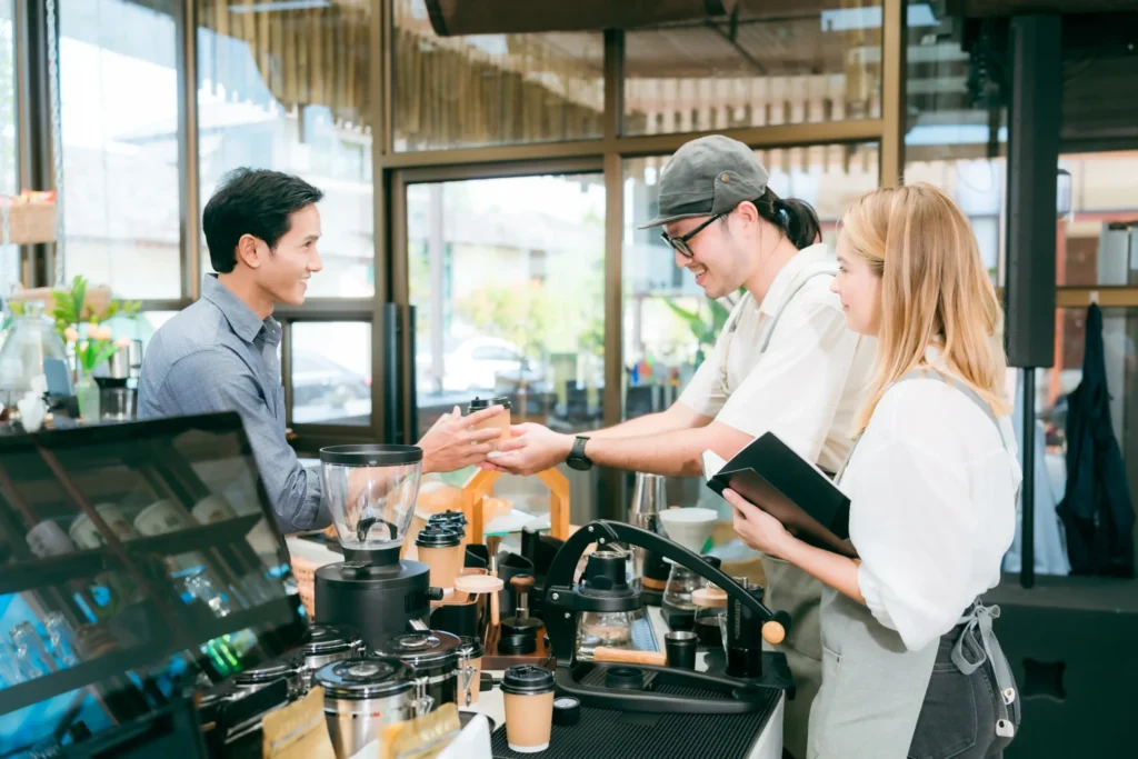 Baristas handing coffee to a smiling guest inside a bright, modern coffee shop, representing teamwork and hospitality in cafe ideas to attract customers.