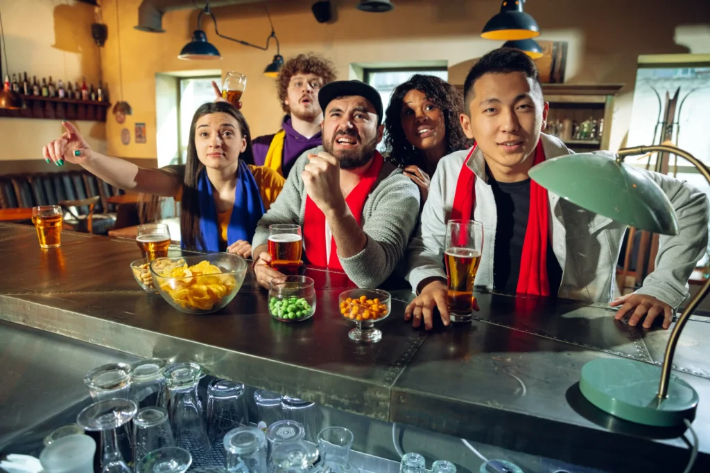 Diverse group of sports fans cheering at a pub while drinking beer, showing community engagement in social media marketing for bars.