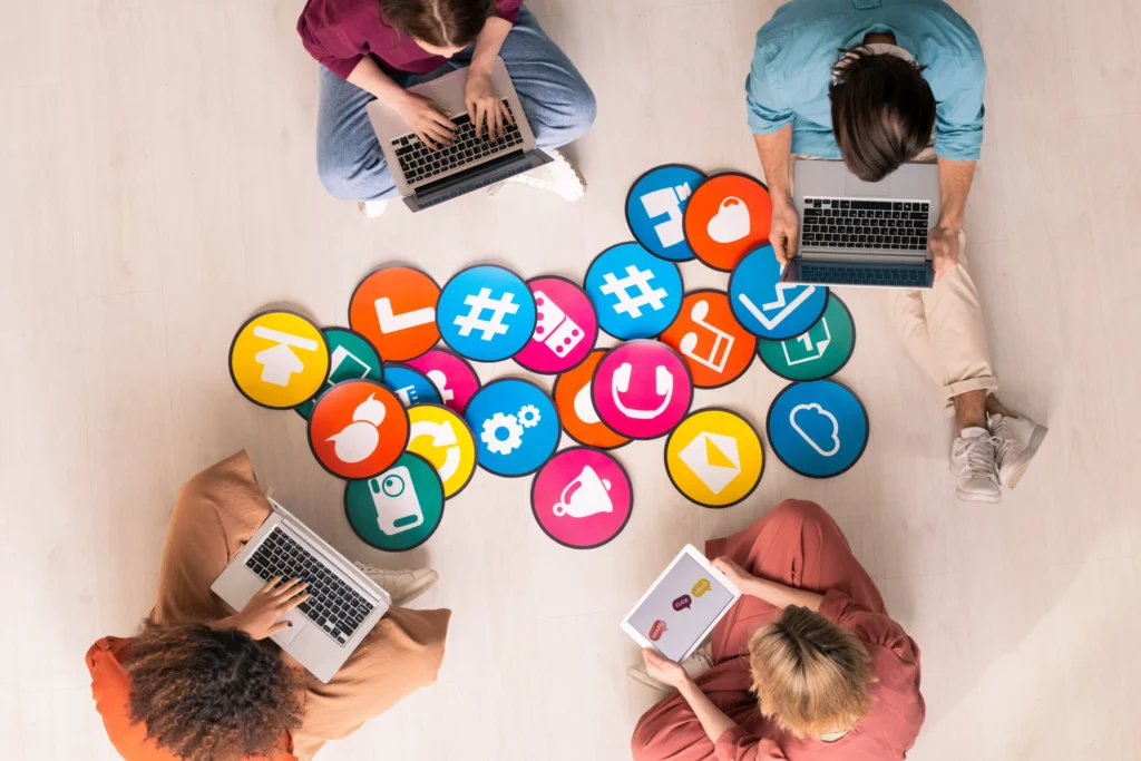 Group of people sitting around colorful social media icons while using laptops and tablets, highlighting social media’s role in a digital marketing strategy for restaurants.