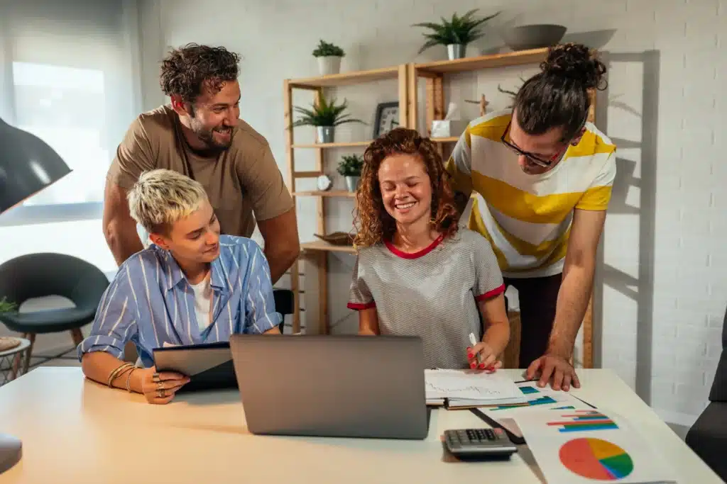 A creative team collaborating around a laptop in a digital web agency, reviewing charts and discussing marketing strategies.