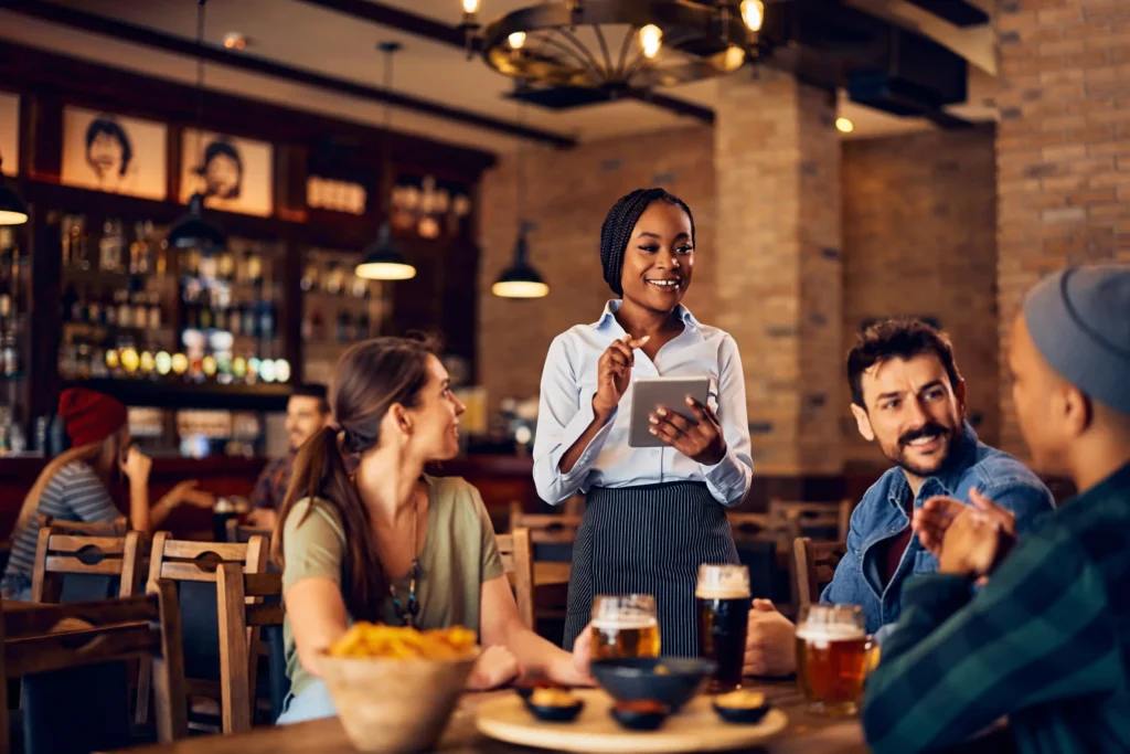 A smiling waitress taking orders with a tablet inside a busy restaurant, highlighting restaurant advertising ideas using technology and service quality.